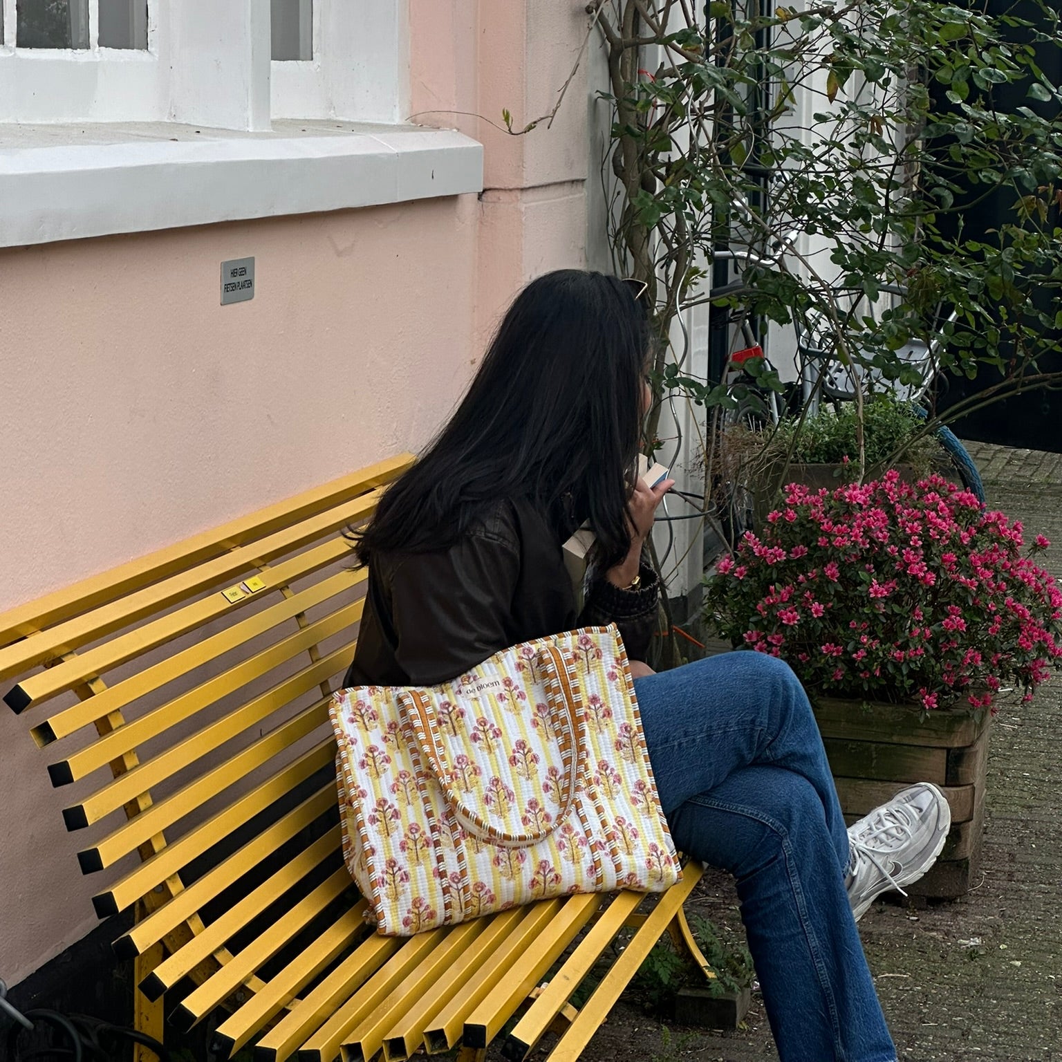 Person sitting on a yellow bench with a floral bag, surrounded by plants and flowers.