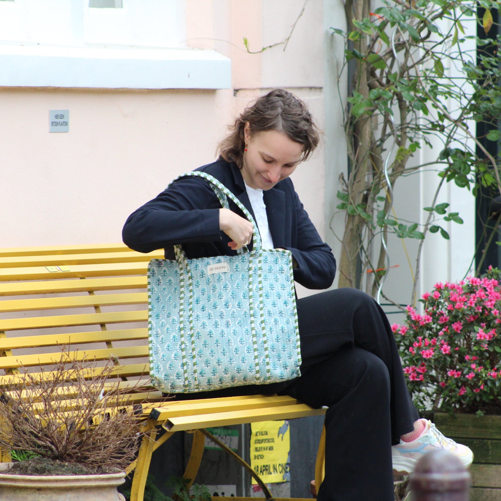 Woman sitting on a bench looking into her handbag with a yellow bench and potted plants in the background.