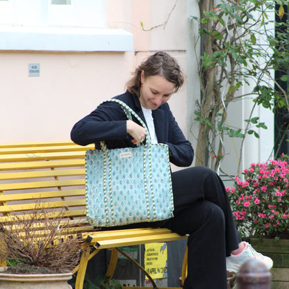 Woman sitting on a bench looking into her handbag with a yellow bench and potted plants in the background.
