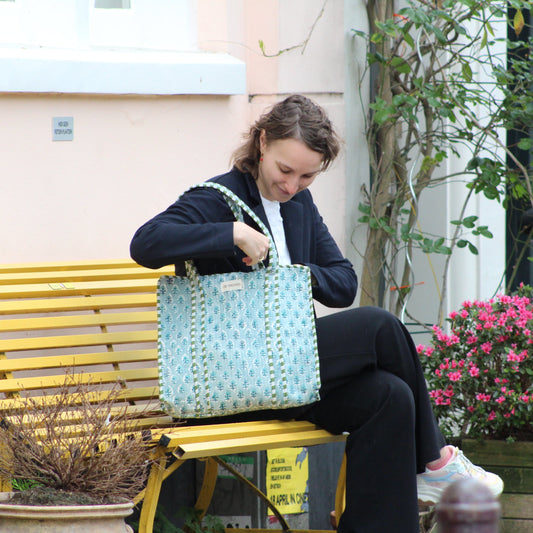 Woman sitting on a bench looking into her handbag with a yellow bench and potted plants in the background.