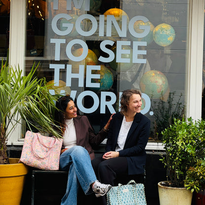 Two women sitting outside a store with a window display and plants.