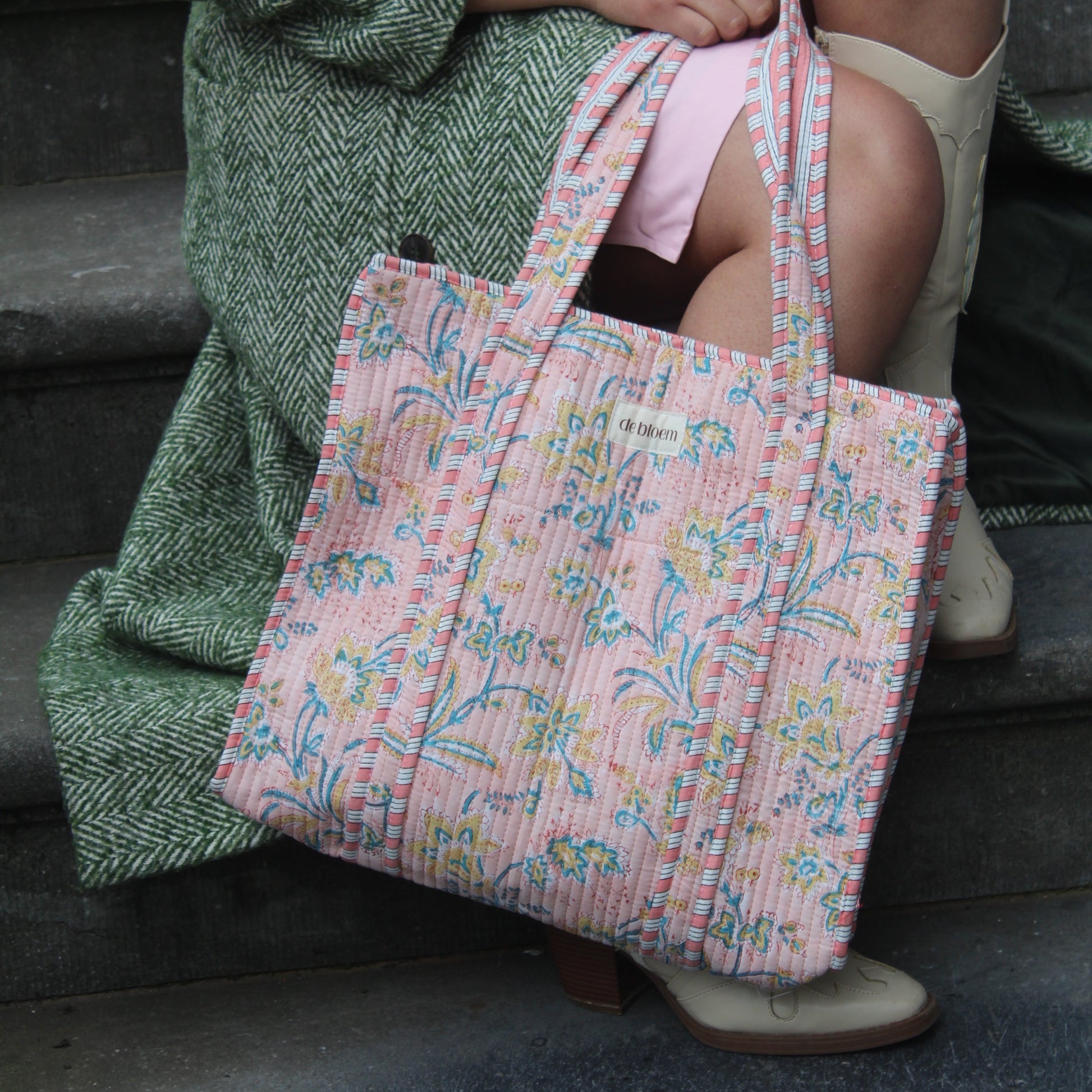 Person holding a pink patterned bag with a green textured blanket draped over them, sitting against a dark background.