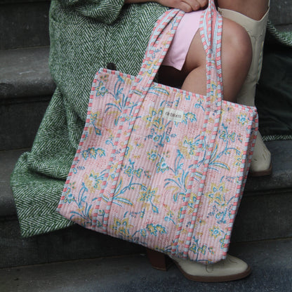 Person holding a pink patterned bag with a green textured blanket draped over them, sitting against a dark background.