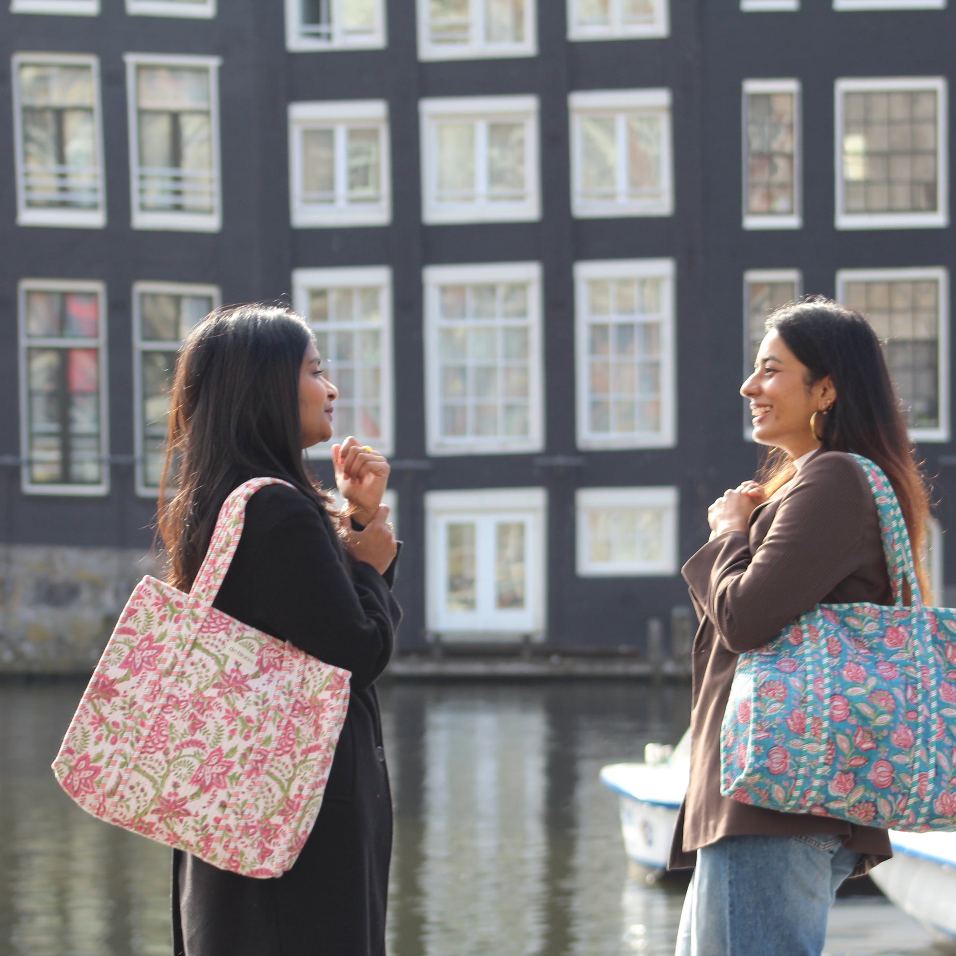 Two women standing by a canal with floral-patterned bags, buildings in the background